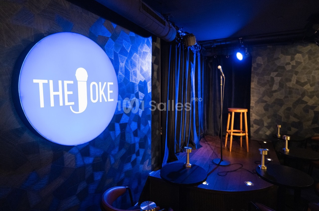 Salle de stand-up avec un micro sur scène, un tabouret en bois et un panneau lumineux indiquant ’The Joke’.