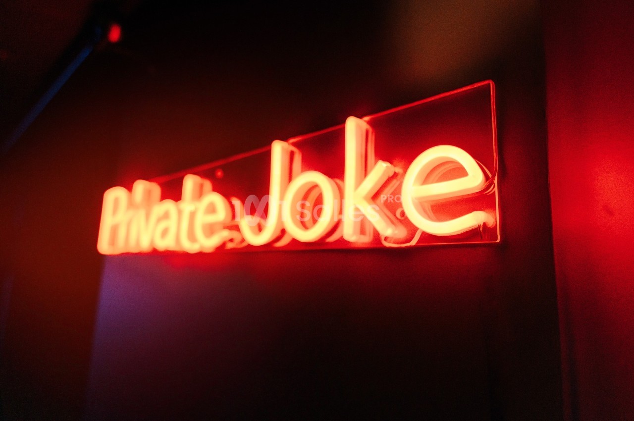 Enseigne lumineuse rouge avec l'inscription ’Private Joke’ dans un environnement sombre.