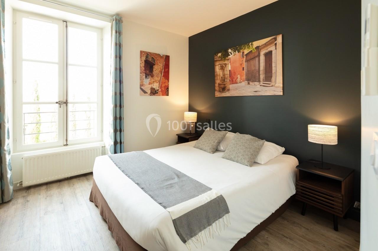 Chambre lumineuse avec un grand lit, murs décorés de tableaux, lampes de chevet et fenêtre avec rideaux à motifs.
