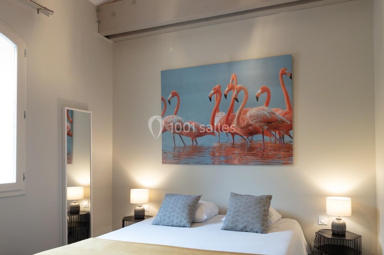 Chambre avec lit double, plaid jaune, tableau de flamants roses, miroir et éclairage suspendu sous plafond en bois.