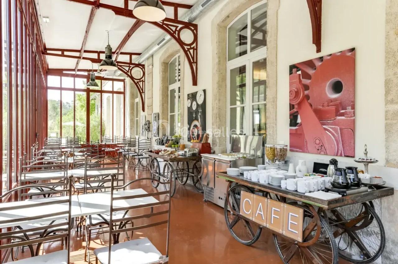 Salle lumineuse avec tables et chaises en métal, buffet de boissons et nourriture, grandes fenêtres et décoration…