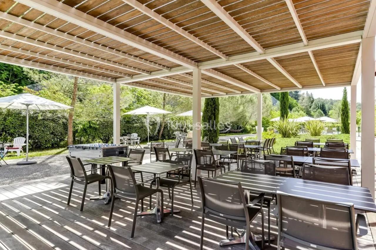 Terrasse extérieure avec tables et chaises sous une pergola en bois, entourée de verdure et de parasols.