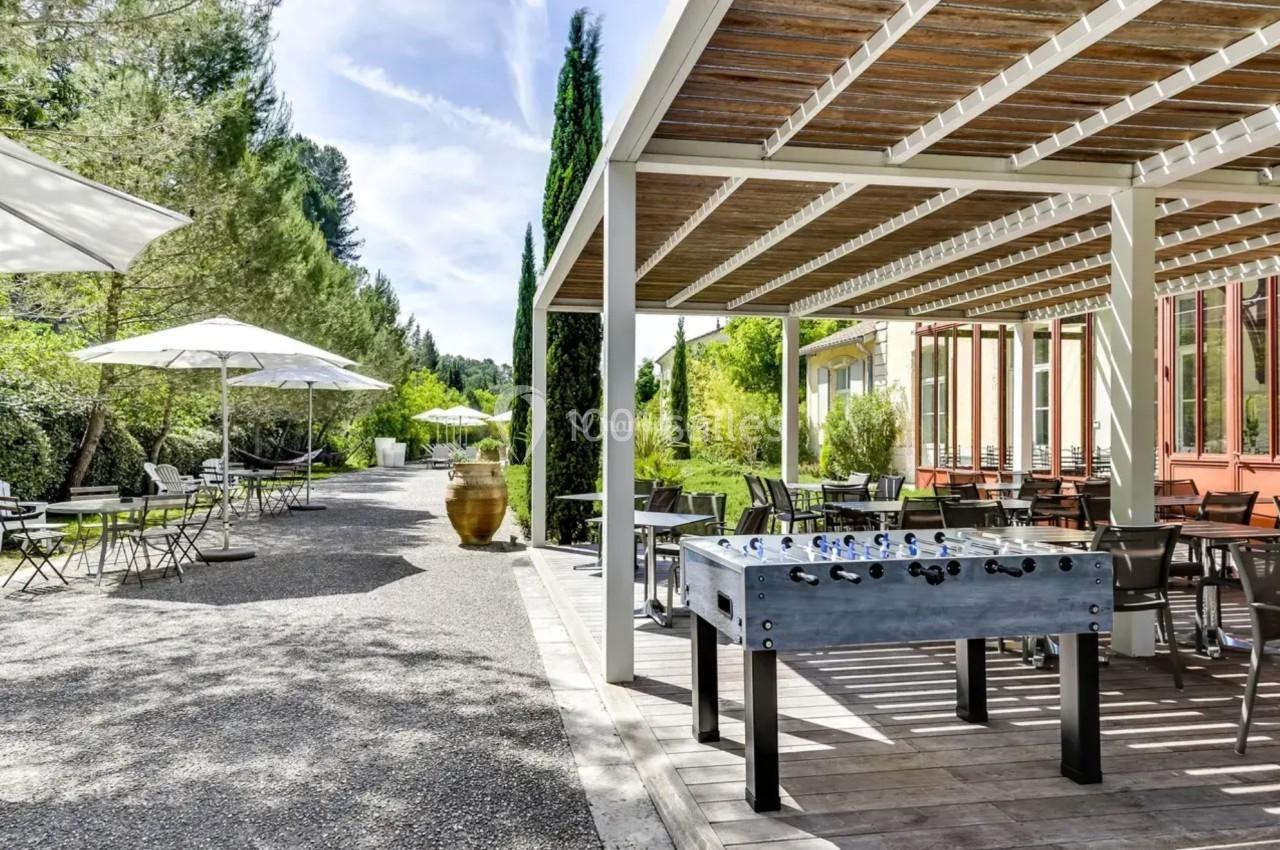 Terrasse extérieure avec pergola, baby-foot, tables, chaises et parasols, entourée de végétation.
