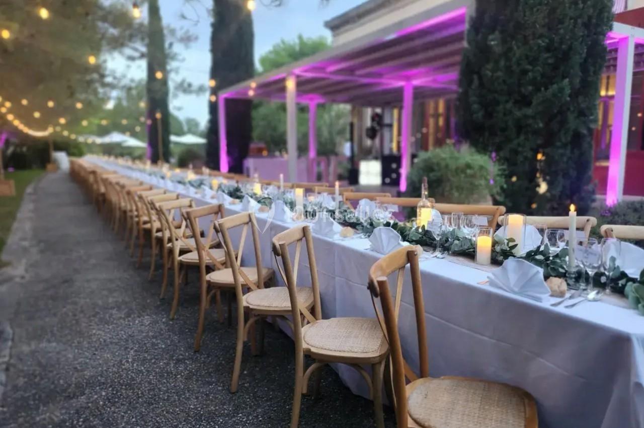 Grande table extérieure décorée pour un dîner, avec nappes blanches, chaises en bois et éclairage suspendu.