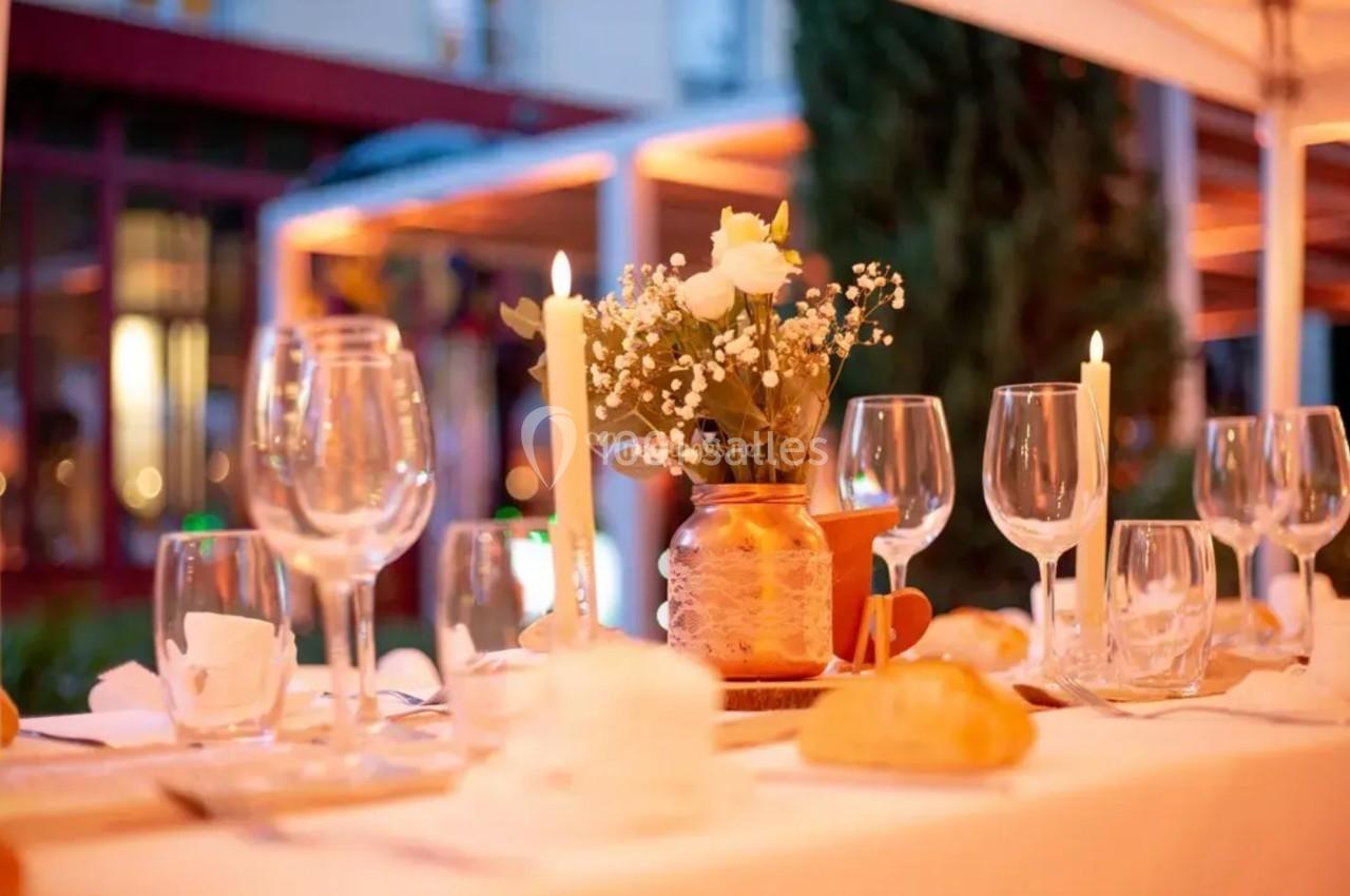 Table dressée avec des verres, bougies allumées et un bouquet de fleurs blanches dans un vase, en extérieur.
