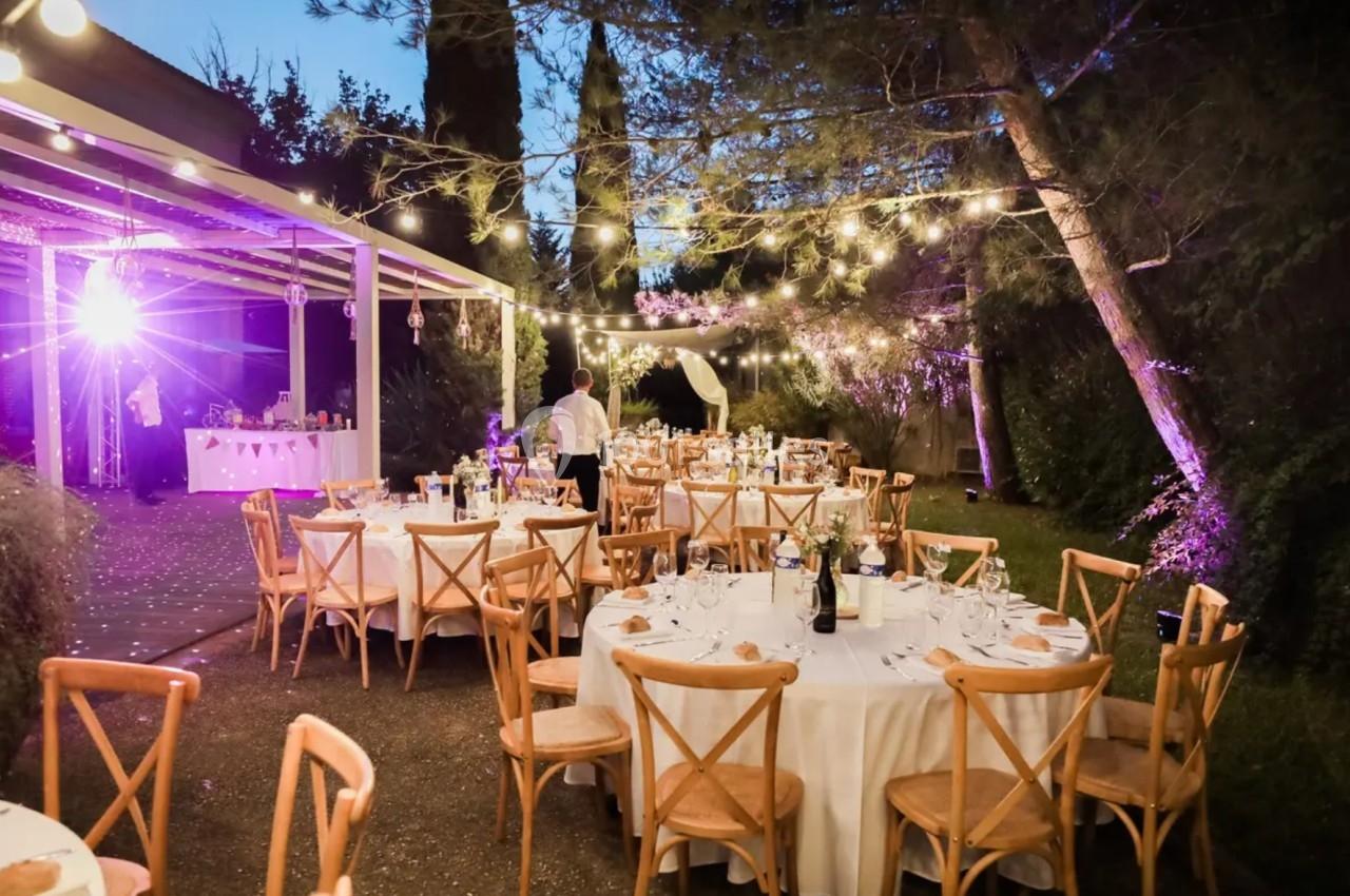 Tables rondes décorées pour un dîner en extérieur, éclairées par des guirlandes lumineuses dans un jardin au crépuscule.