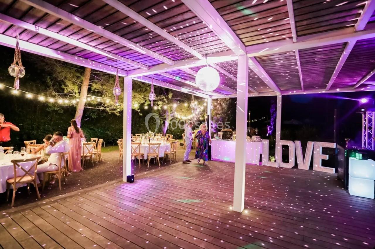 Terrasse en bois éclairée par des guirlandes lumineuses et un éclairage violet, avec des tables dressées et un décor ’LOVE’.