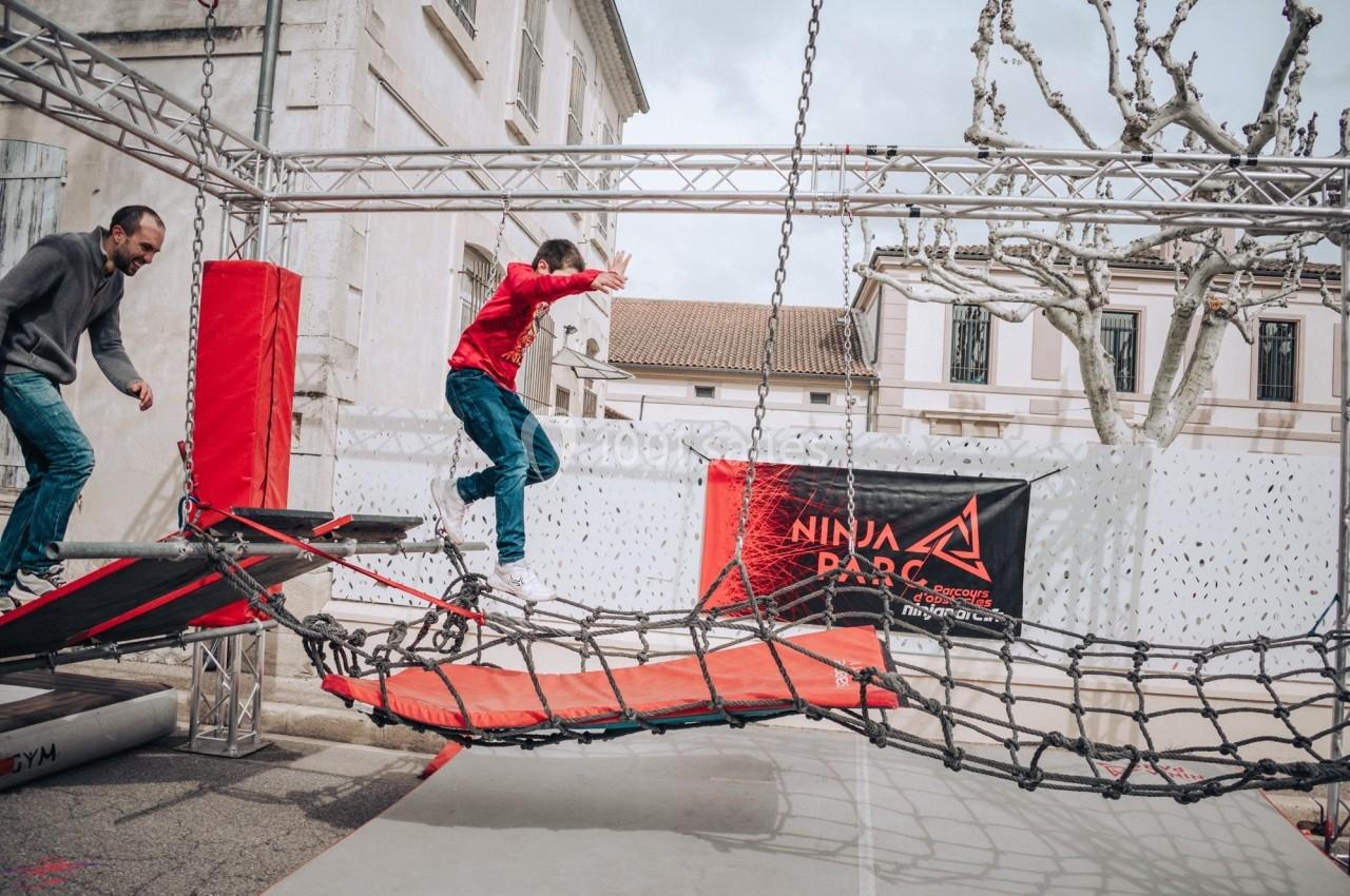 Un enfant traverse un parcours d'obstacles suspendus en extérieur, sous la supervision d'un adulte.