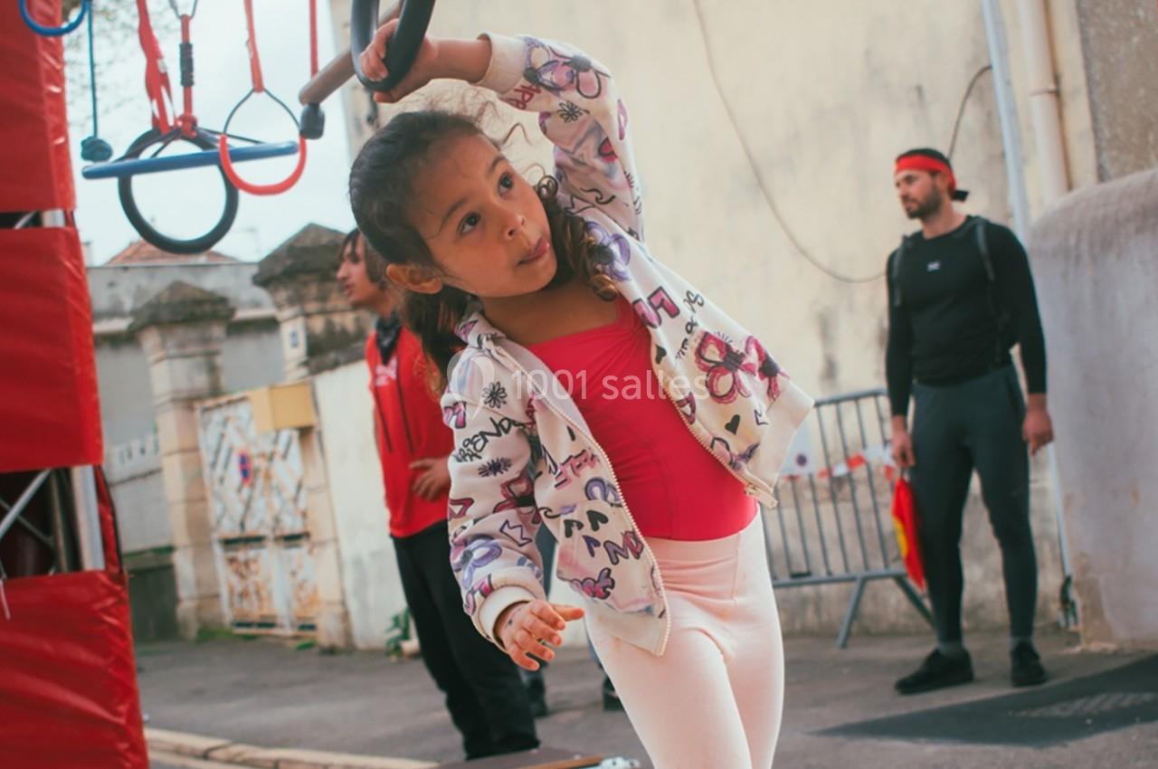 Une fillette en tenue de sport s'entraîne sur un parcours d'obstacles en extérieur sous la supervision d'adultes.