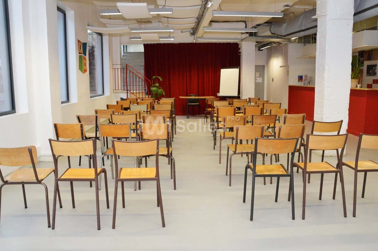 Salle lumineuse avec des rangées de chaises en bois disposées face à un tableau blanc et un rideau rouge.