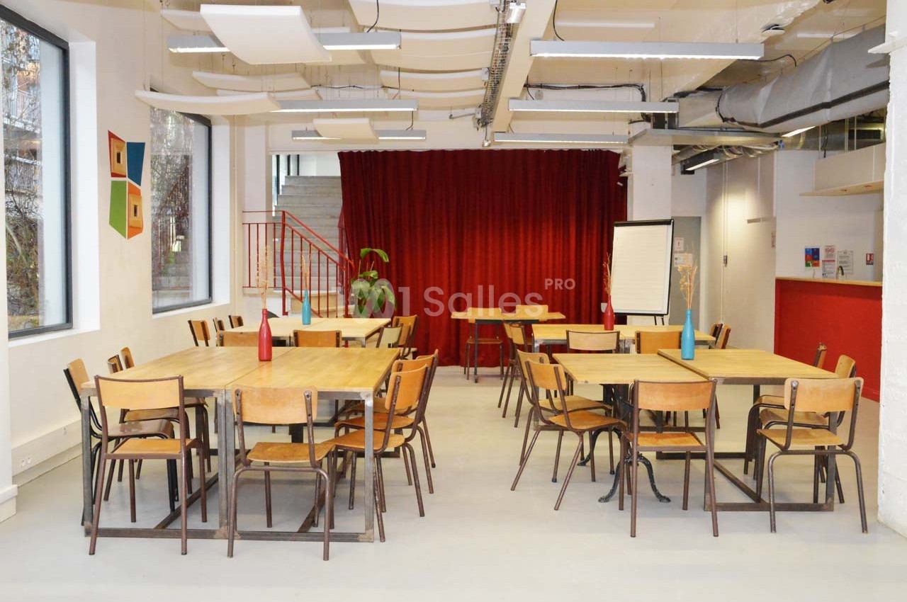 Salle lumineuse avec tables et chaises en bois, rideau rouge en fond, plantes et tableau blanc sur le côté.