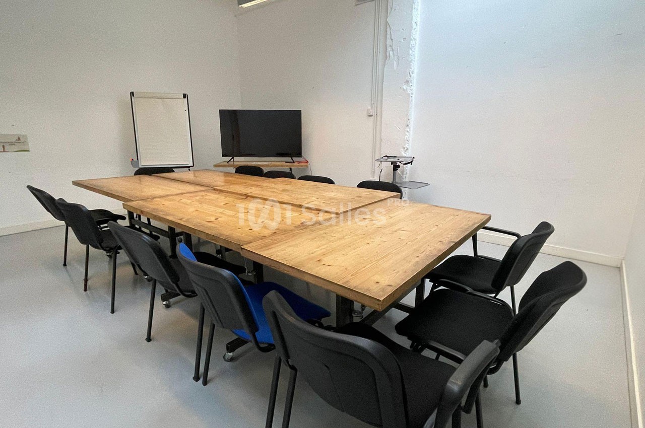 Salle de réunion avec une grande table en bois entourée de chaises noires, un écran et un tableau blanc en arrière-plan.