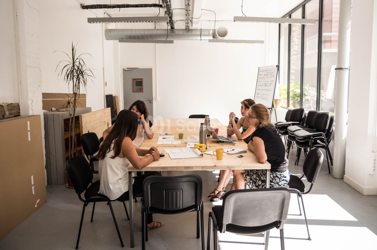 Quatre personnes discutent autour d'une table dans une salle de réunion lumineuse avec des fenêtres et des chaises.