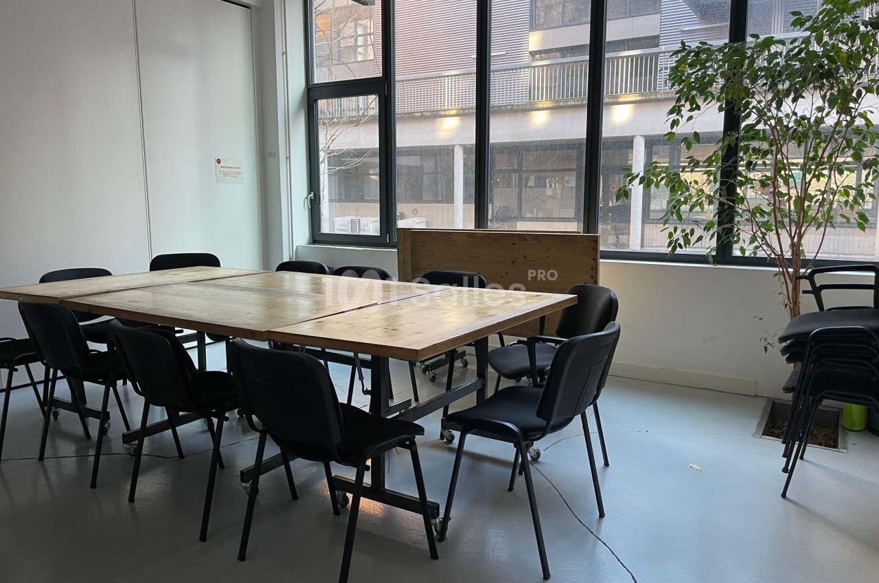 Salle de réunion lumineuse avec une grande table en bois entourée de chaises noires, près de fenêtres et d'une plante.