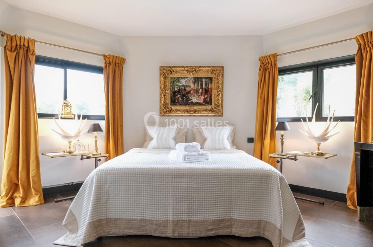 Chambre lumineuse avec lit double, draps blancs, rideaux jaunes, tableaux encadrés et fenêtres sur les côtés.