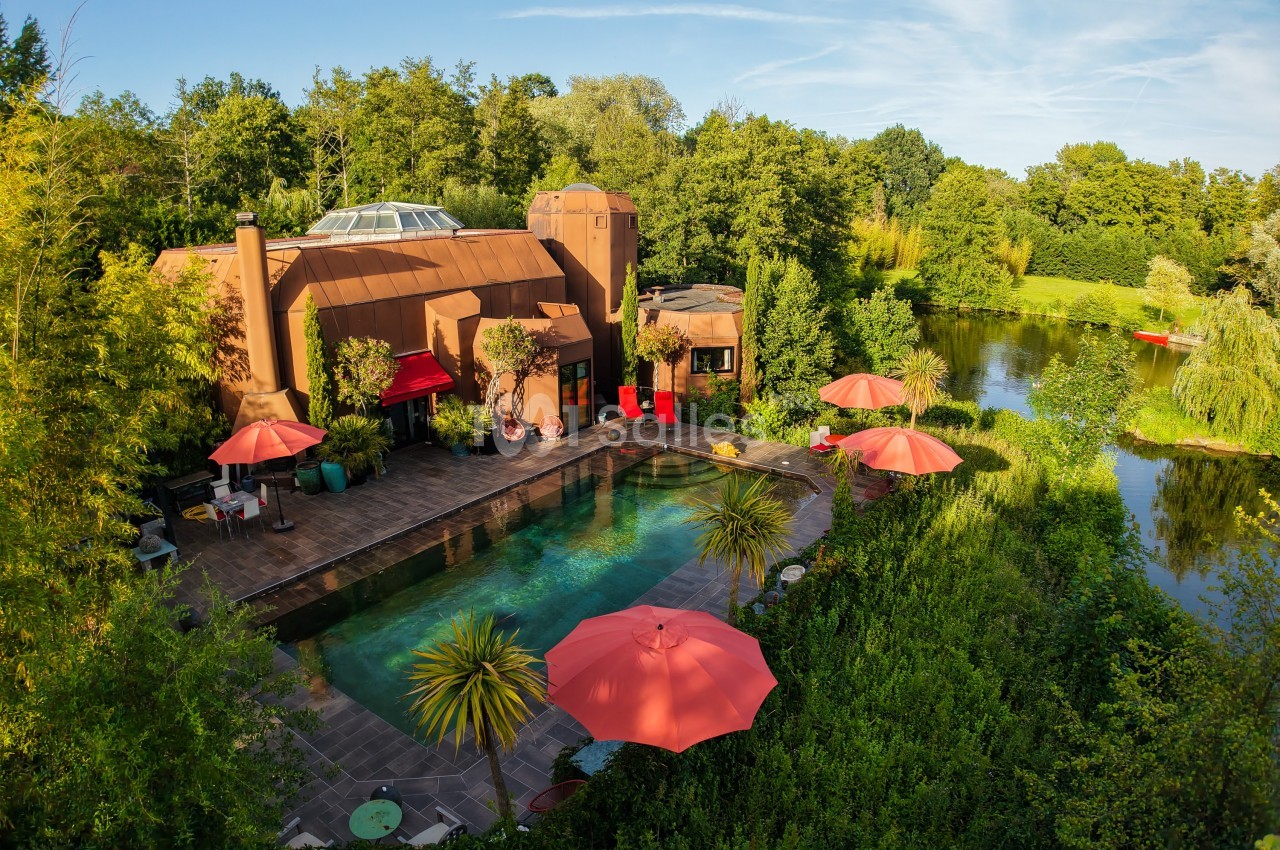 Vue aérienne d'une maison moderne entourée de verdure, avec piscine, terrasses et parasols rouges près d'un étang.