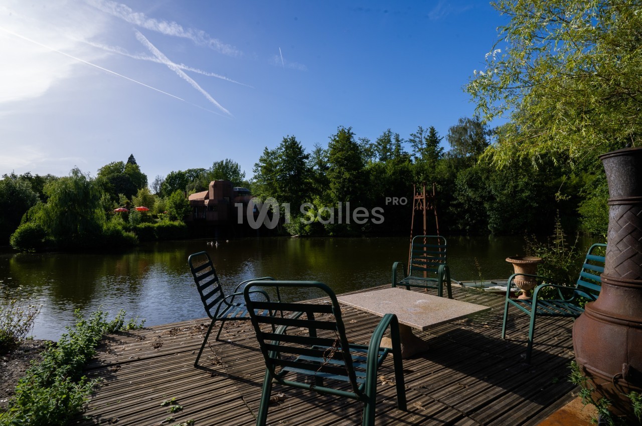 Terrasse en bois avec des chaises autour d'une table, donnant sur un étang entouré de végétation et d'arbres.