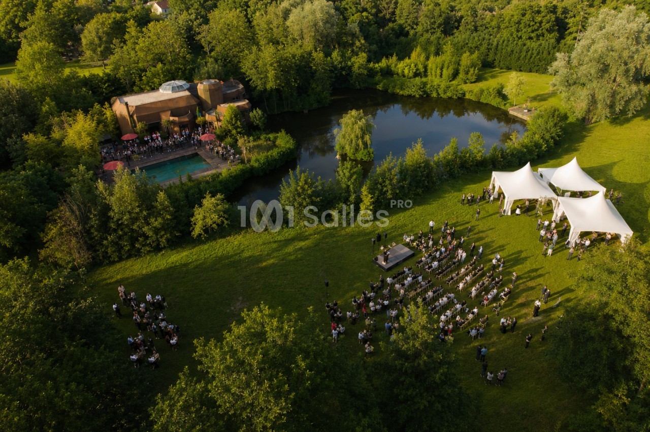 Vue aérienne d'un événement en plein air avec invités, tentes blanches, scène et étang entouré de verdure.