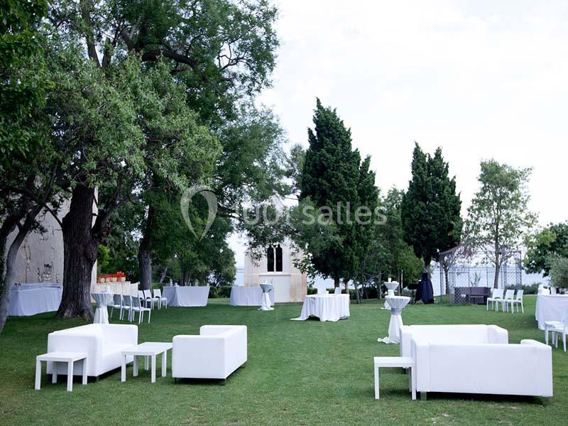 Espace extérieur avec pelouse, mobilier blanc moderne et tables dressées, entouré d'arbres et de verdure.