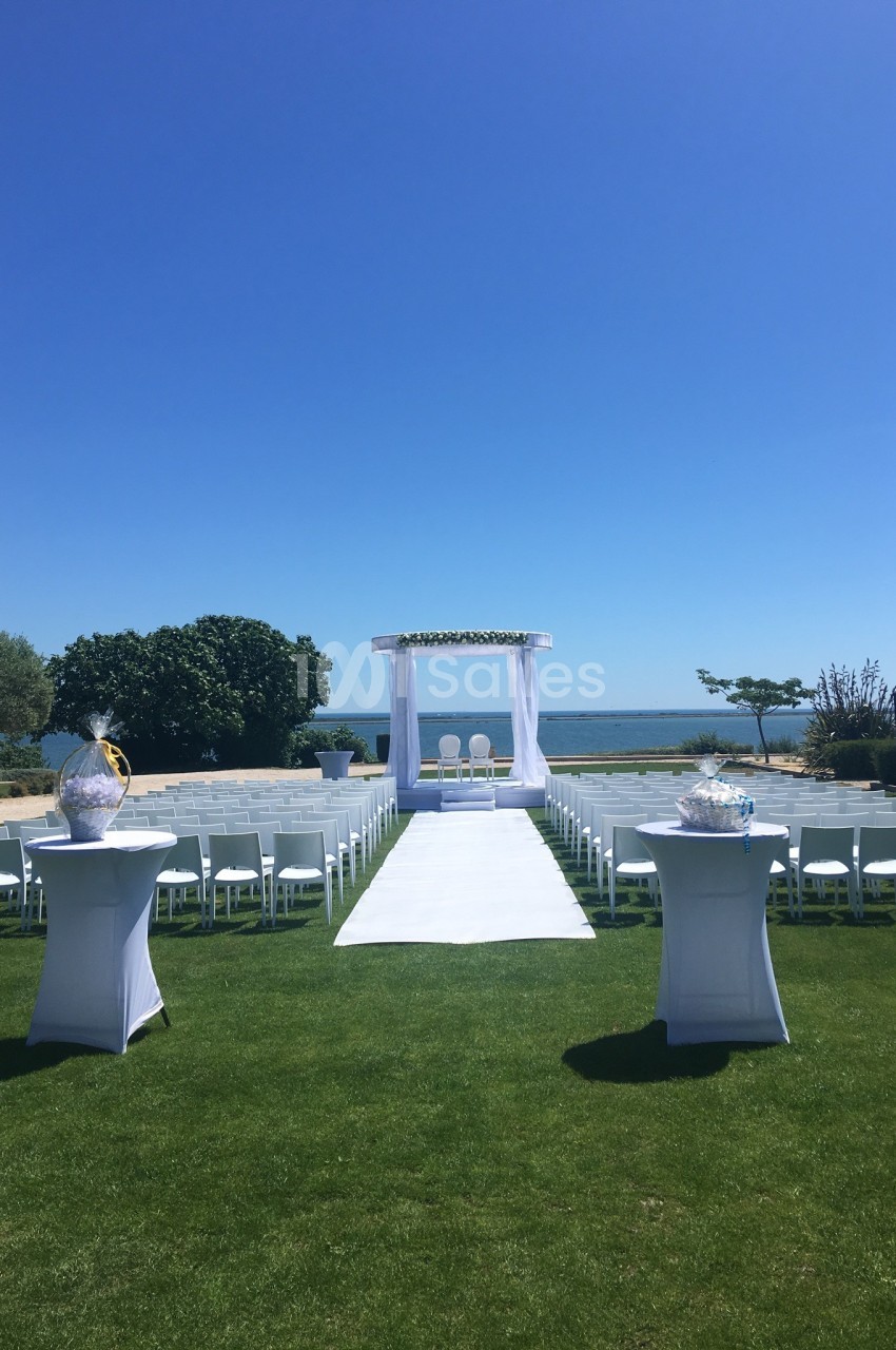 Allée centrale menant à une arche de mariage blanche, entourée de chaises, avec vue sur la mer et un ciel dégagé.