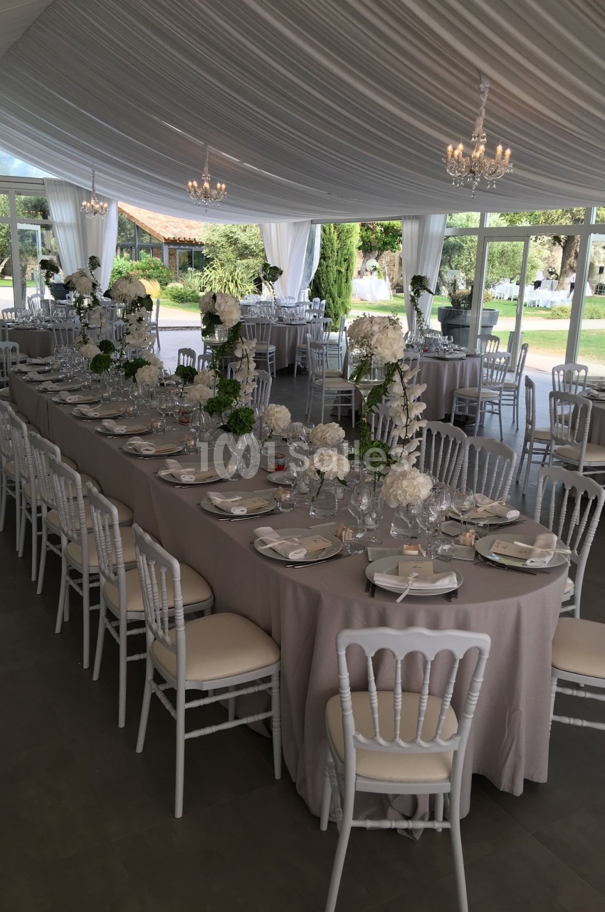 Salle de réception élégante avec tables dressées, nappes beiges, chaises blanches et décor floral sous un plafond drapé.