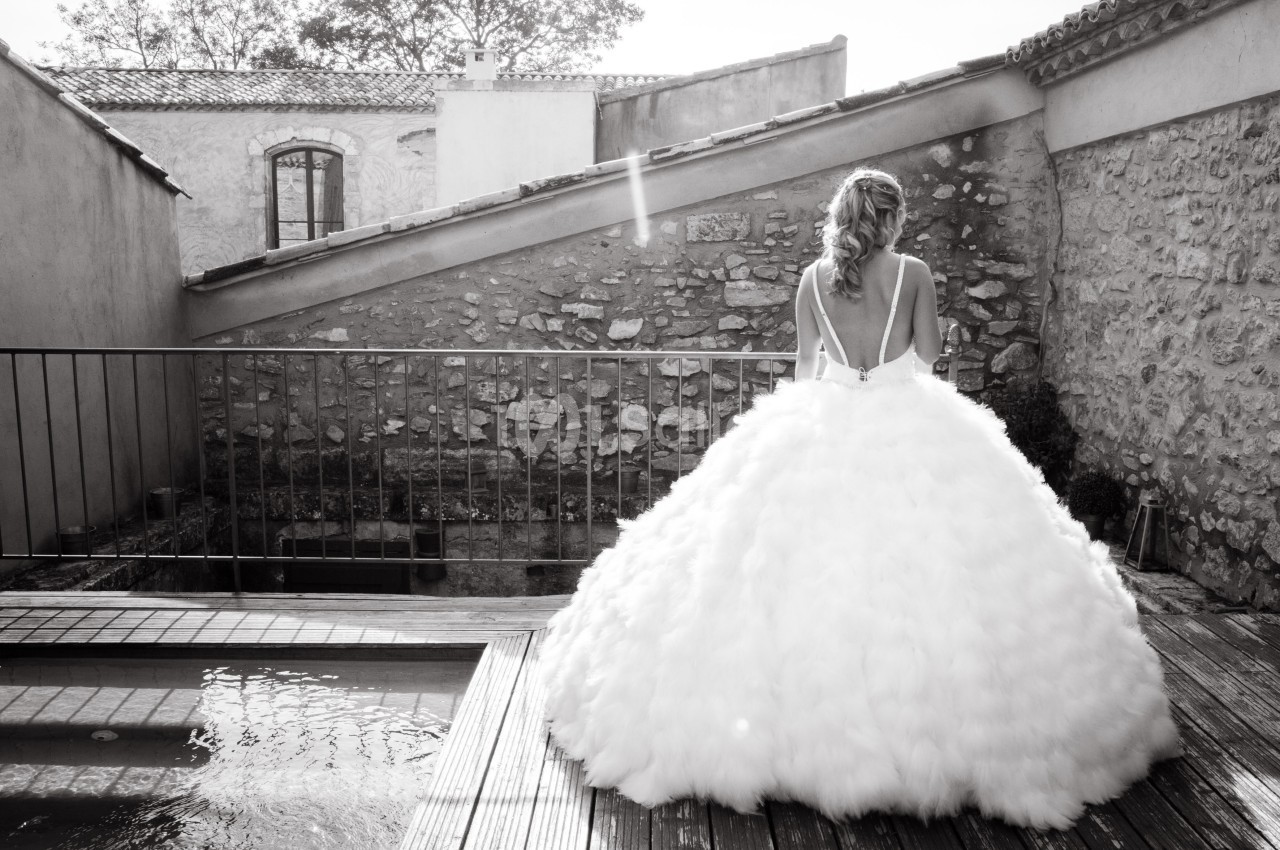 Une femme en robe de mariée volumineuse vue de dos, debout sur une terrasse en bois près d'une petite piscine.
