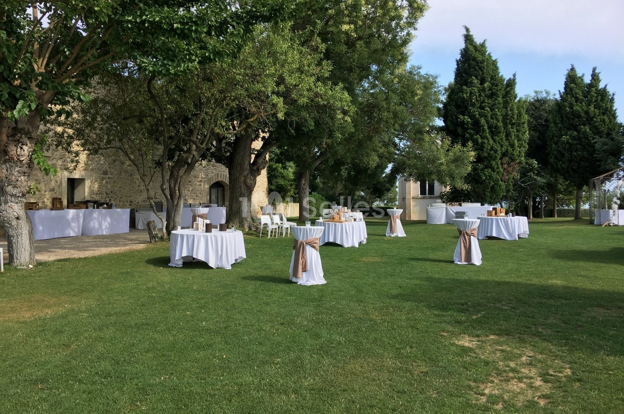 Tables rondes dressées avec nappes blanches et décorations beige, disposées sur une pelouse entourée d'arbres.