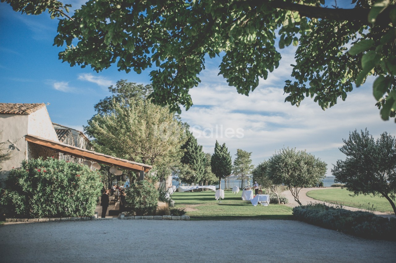 Jardin extérieur avec pelouse, arbres, tables dressées et bâtiment en pierre sous un ciel bleu clair.