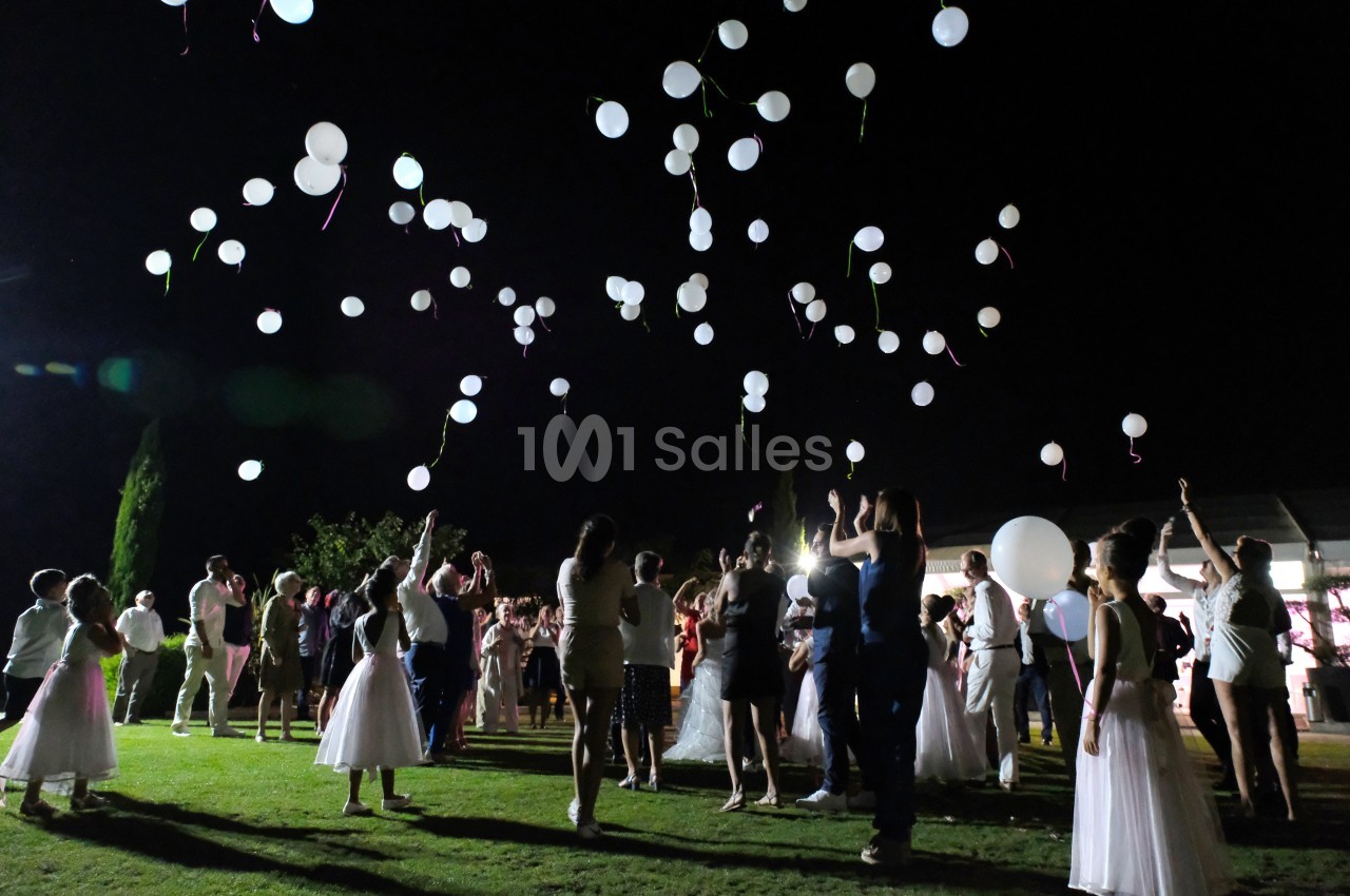Des personnes en tenue de soirée lâchent des ballons blancs dans un jardin éclairé la nuit.