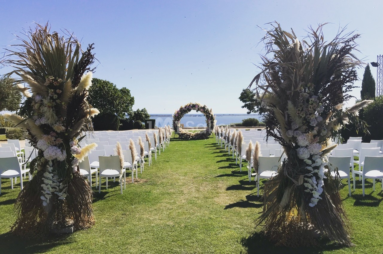 Allée centrale décorée de fleurs séchées menant à une arche florale sur une pelouse, avec vue sur la mer.