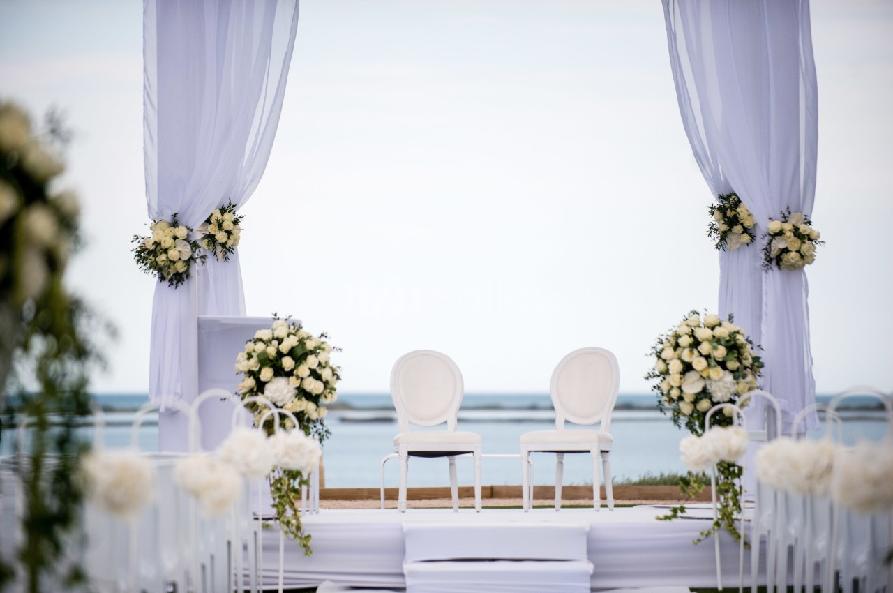 Deux chaises blanches sous une arche décorée de fleurs blanches, face à la mer dans un cadre extérieur.
