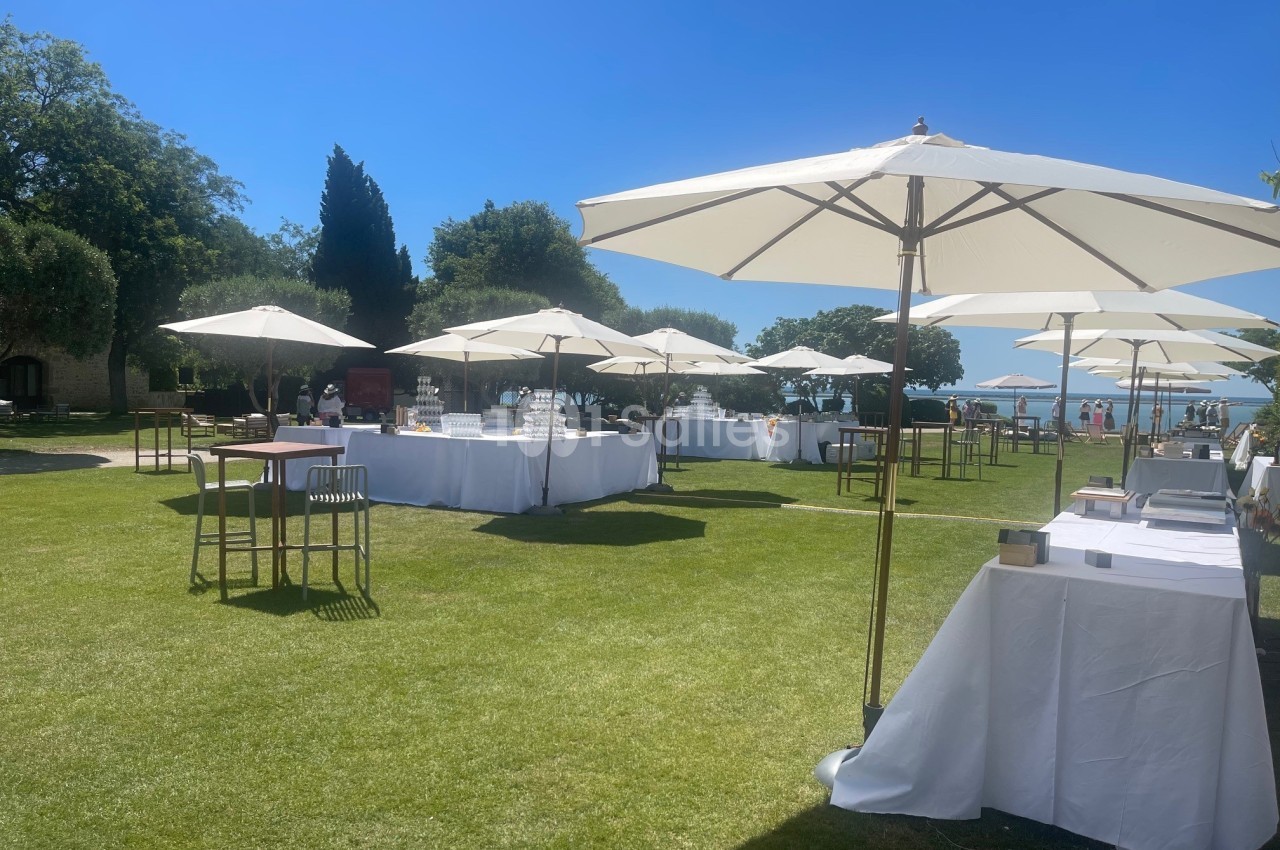 Tables dressées sous des parasols blancs sur une pelouse ensoleillée, entourée d'arbres et d'un ciel bleu.