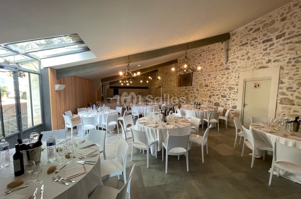 Salle de réception lumineuse avec murs en pierre, tables rondes dressées pour un repas et grandes baies vitrées.