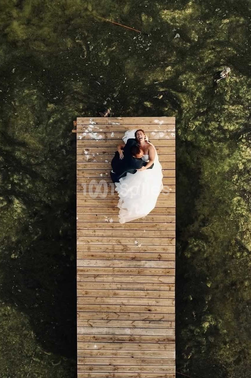 Un couple en tenue de mariage assis sur un ponton en bois au-dessus d'une eau verdâtre vue d'en haut.