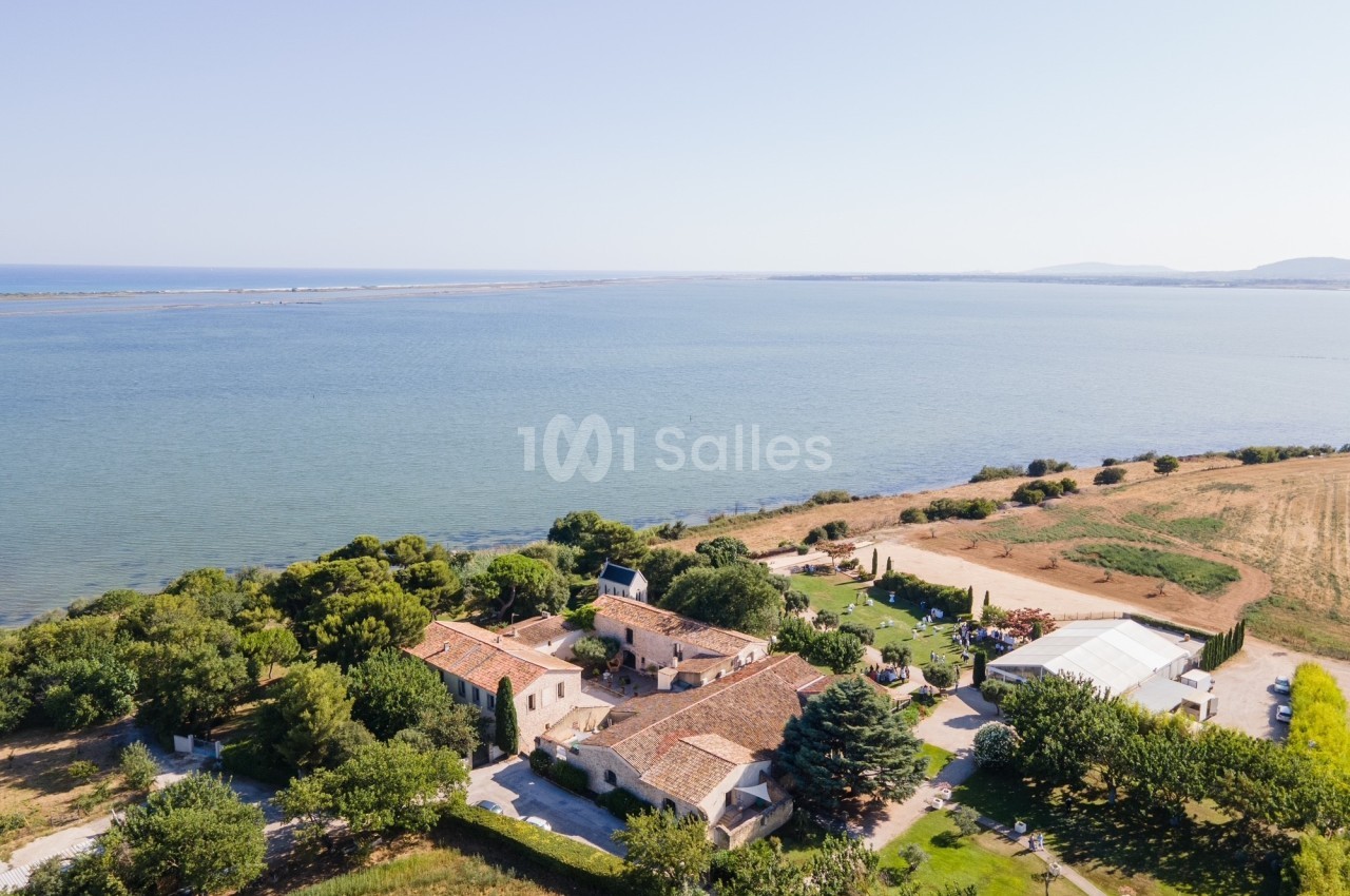 Vue aérienne d'un domaine rural avec bâtiments en pierre, arbres et vue sur une étendue d'eau calme en arrière-plan.