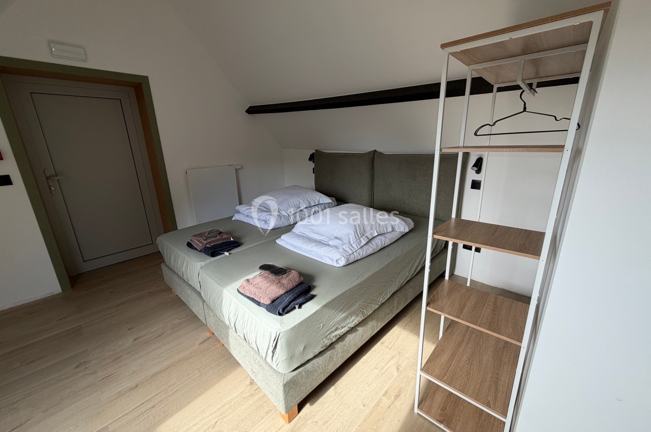 Chambre avec lit double, linge plié sur le lit, étagère en bois et métal, sol en parquet clair.