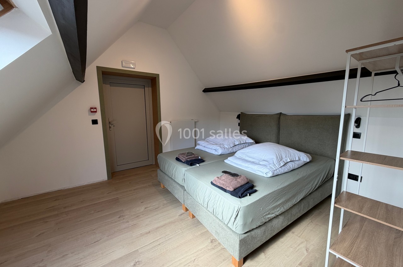 Chambre avec deux lits simples rapprochés, étagère ouverte et plafond mansardé dans un intérieur lumineux.