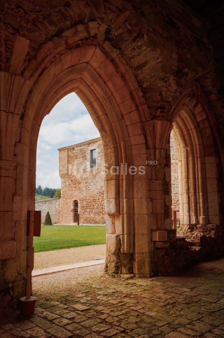 Vue depuis des arcs en pierre d'une cour herbeuse entourée de bâtiments anciens en pierre sous un ciel partiellement nuageux.