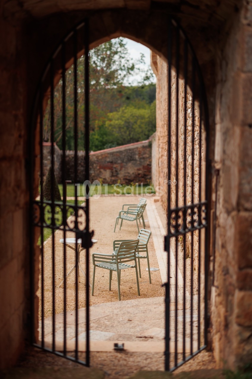 Vue à travers une porte en fer forgé ouverte sur une terrasse avec des chaises en métal et un jardin en arrière-plan.