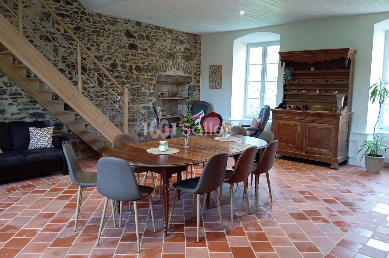 Salle à manger lumineuse avec table en bois, chaises modernes, escalier en bois, murs en pierre et carrelage au sol.