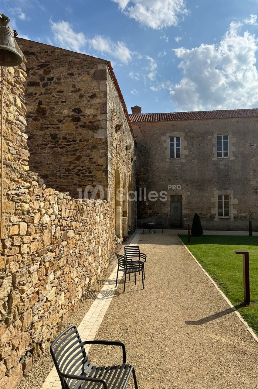 Allée pavée bordée de bancs en métal, longeant un bâtiment en pierre sous un ciel partiellement nuageux.