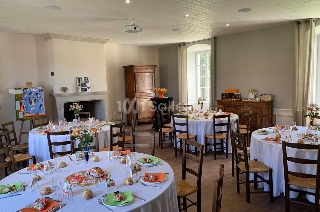 Salle décorée pour un repas festif avec des tables rondes dressées, des chaises en bois et une lumière naturelle.