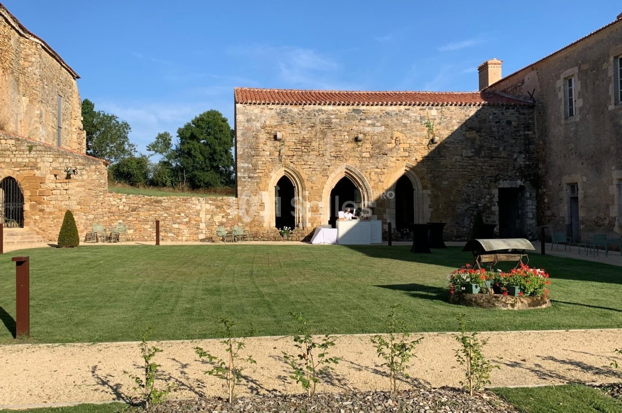 Cour intérieure d'un bâtiment en pierre avec pelouse, parterre fleuri, et tables dressées sous un ciel dégagé.