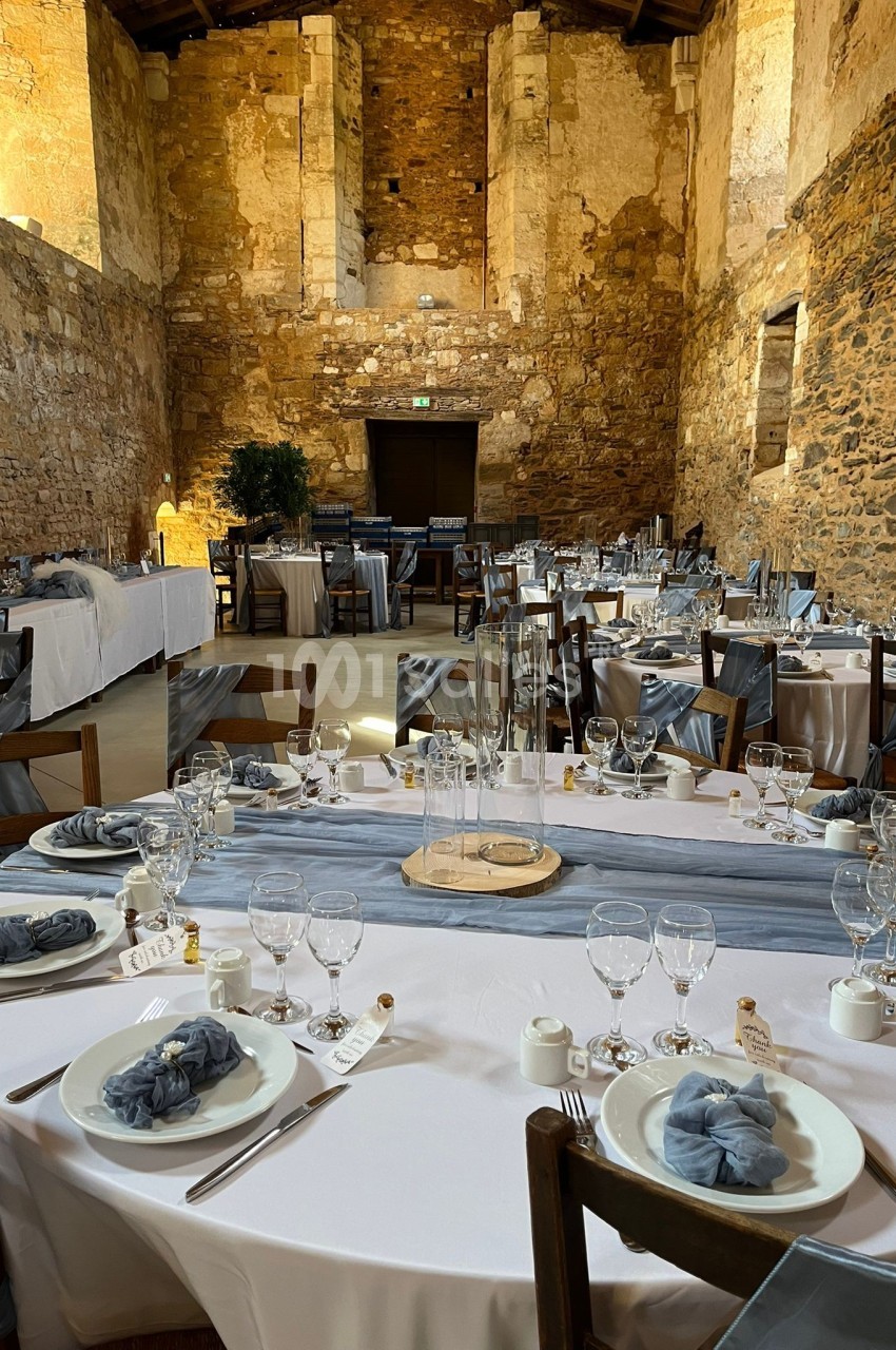 Salle en pierre ancienne aménagée pour un repas, avec tables dressées de nappes blanches et décorations élégantes.
