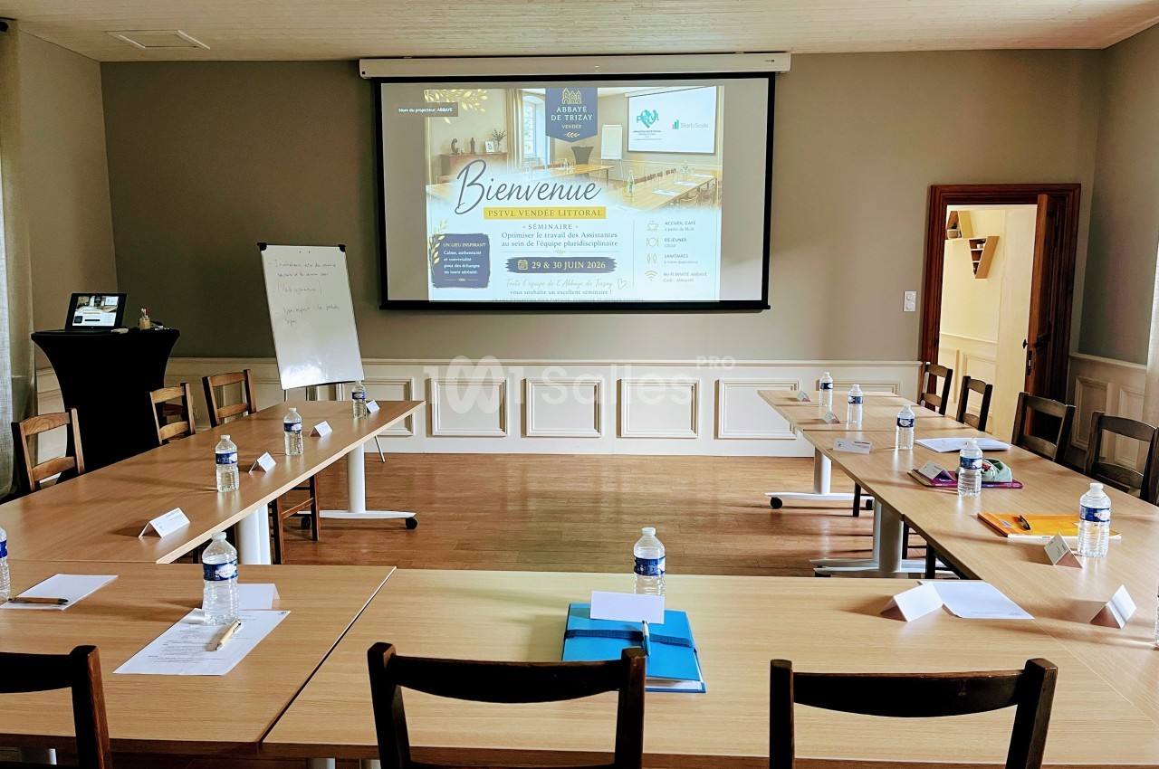 Salle de réunion lumineuse avec tables disposées en U, écran de projection allumé et matériel de travail sur les tables.