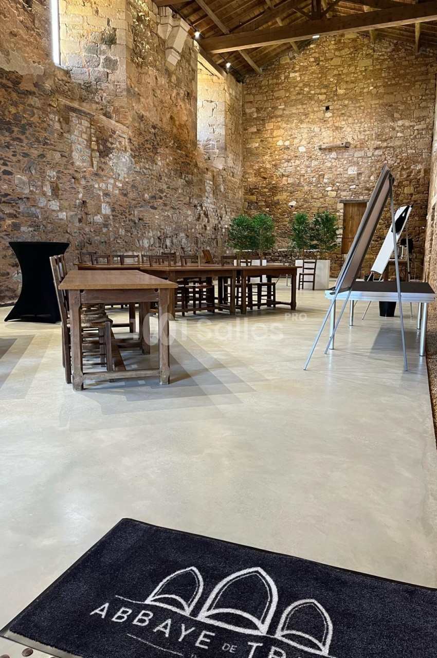 Salle en pierre avec tables en bois, tableau de présentation et tapis marqué ’Abbaye de Trizay’.