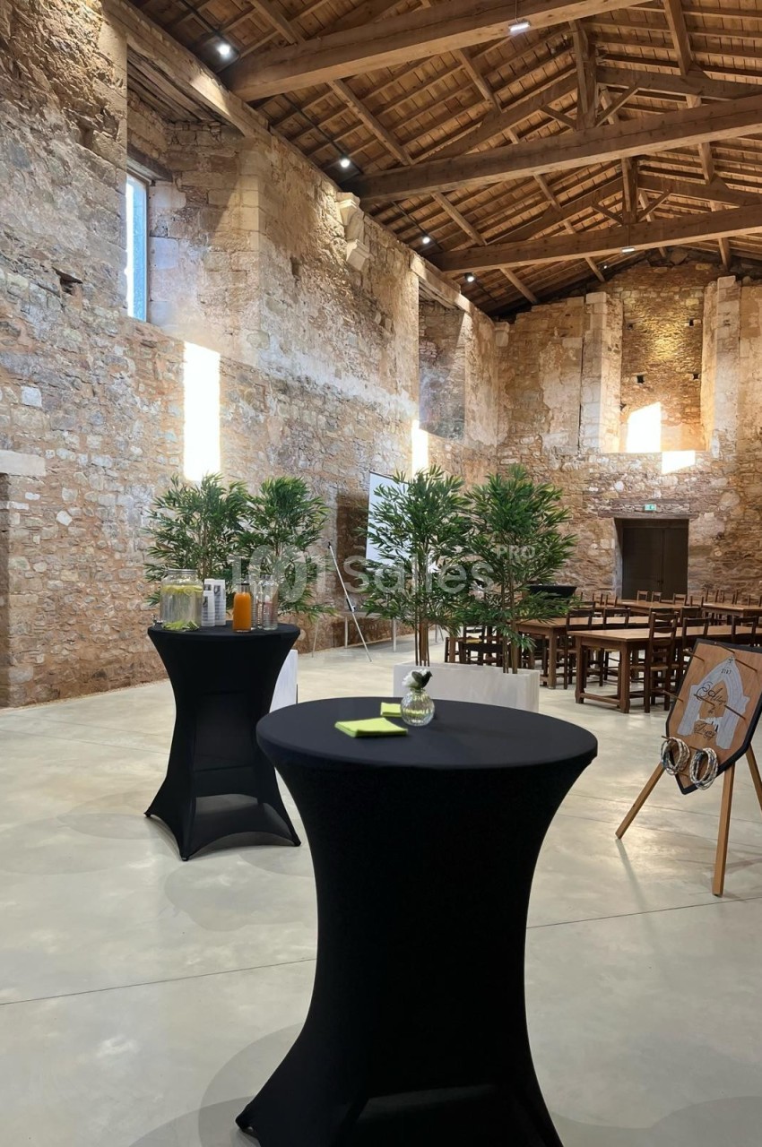 Salle en pierre avec tables hautes noires, boissons, plantes décoratives et chaises disposées pour un événement.