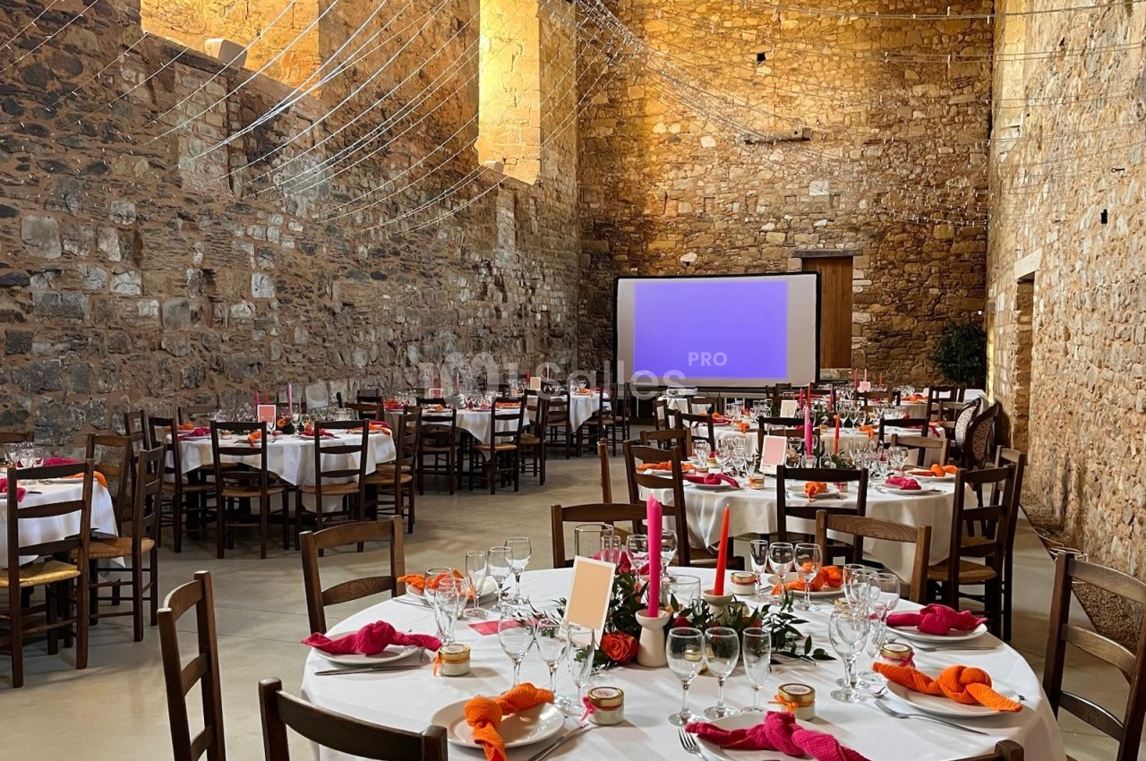 Salle en pierre aménagée pour un événement, avec tables rondes dressées, nappes blanches et décorations colorées.