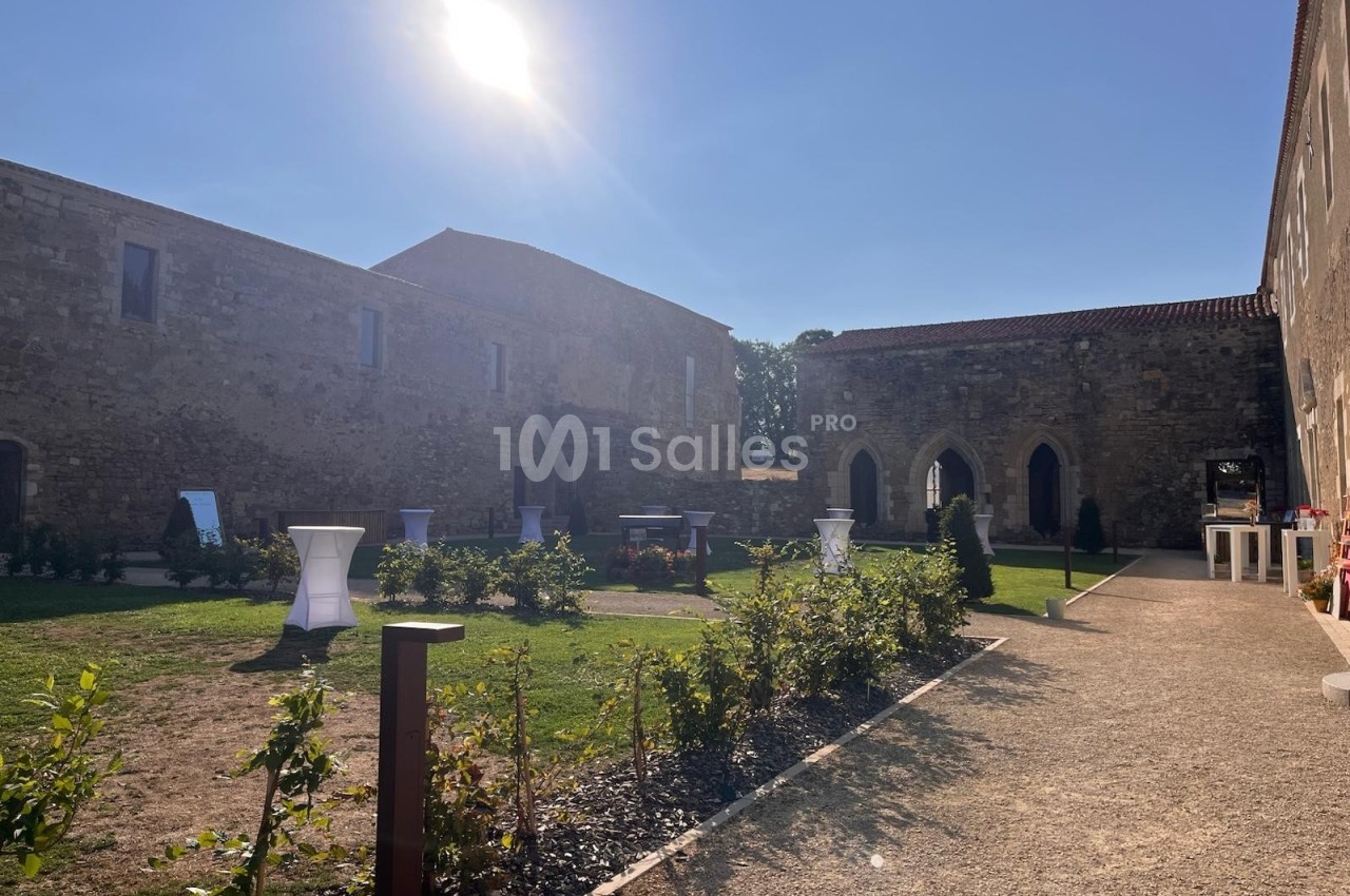 Cour intérieure d'un bâtiment historique en pierre, aménagée avec des tables hautes et éclairée par un soleil éclatant.