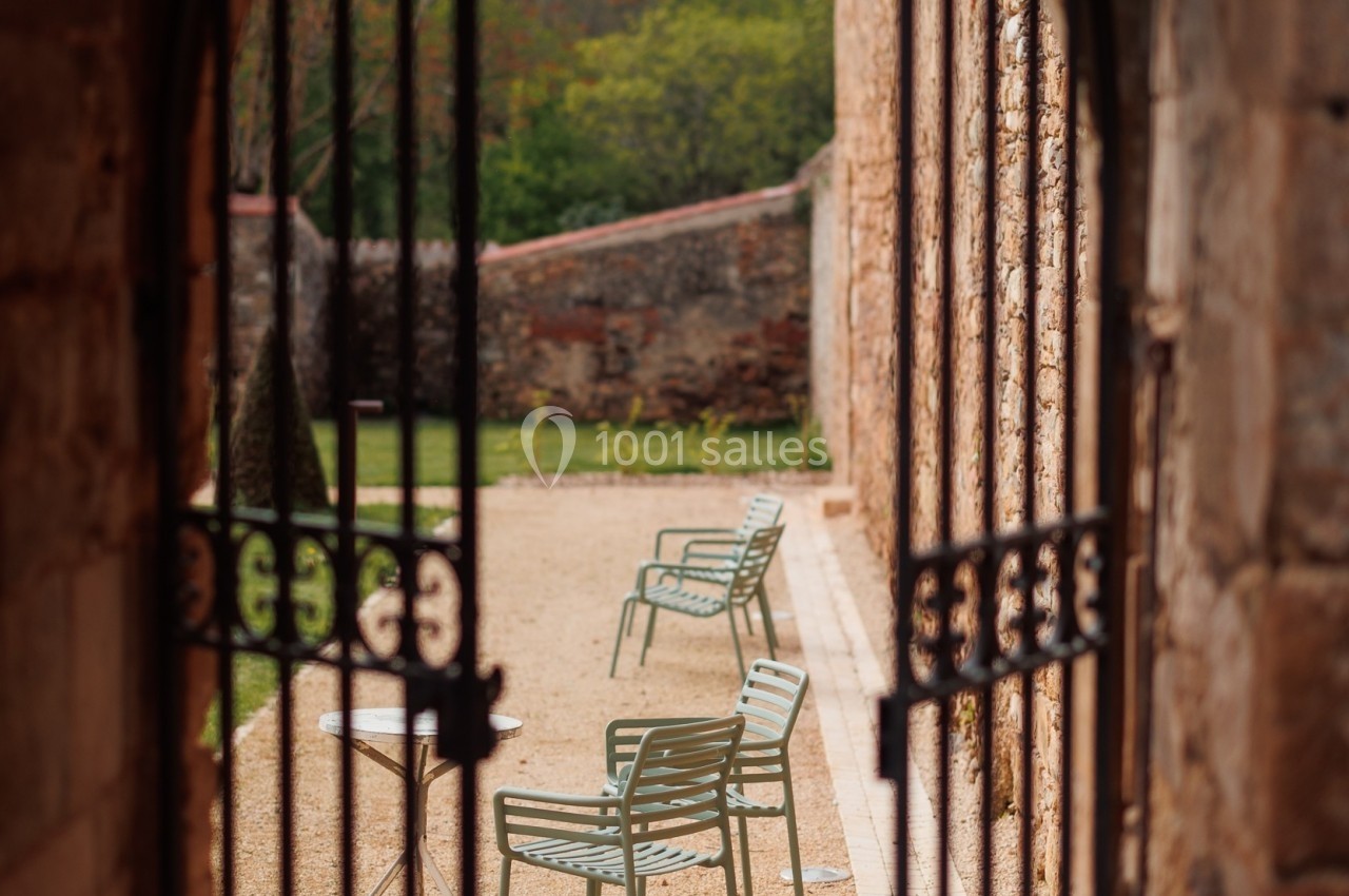 Vue à travers une porte en fer forgé sur des chaises vertes disposées dans une cour en gravier bordée de murs en pierre.
